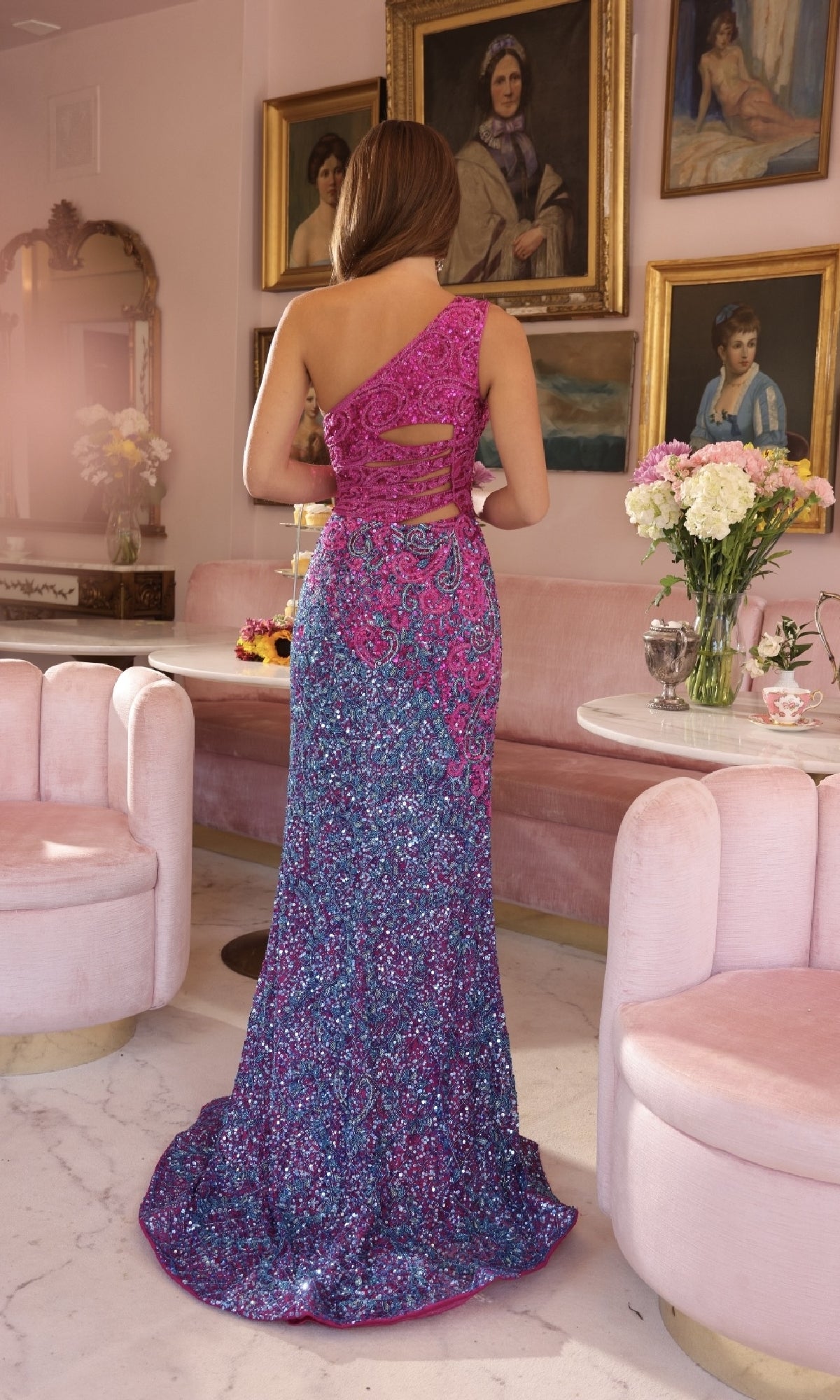 A woman wearing the Ava Presley 47039 long, one-shoulder sequin prom dress stands with her back to the camera in an elegant room featuring pink furniture and floral arrangements.